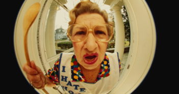 Mature woman standing outside front door, waving spoon (fish-eye)